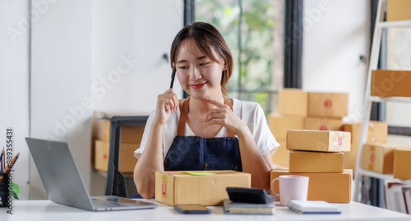 Fototapeta Portrait business woman smile and use laptop checking information on parcel shipping box before send to customer. Entrepreneur small business working at home. SME business online marketing.