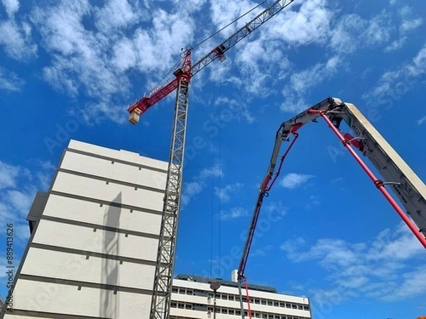 Fototapeta A crane and a building under construction against a blue sky background. Builders work on large construction sites, and there are many cranes working in the field of new construction.
