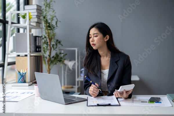 Fototapeta A woman is sitting at a desk with a laptop and a calculator