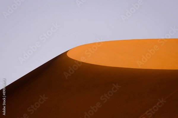 Fototapeta A sunlit sand dune stands out against a clear sky, capturing the minimalist beauty and striking contrasts of the Namibian desert landscape.