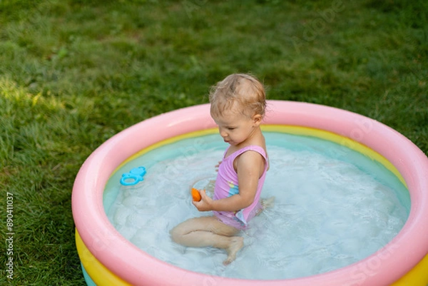 Fototapeta Baby in a pink swimsuit is swimming in an inflatable pool and eating an apricot in the backyard. 