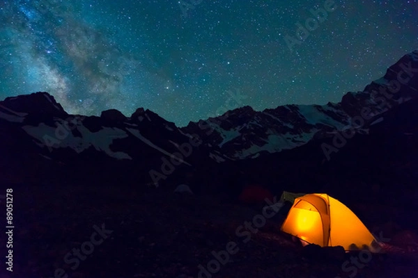 Fototapeta Night mountain landscape with illuminated tent Silhouettes of snowy mountain peaks and edges night sky with many stars and milky way on background illuminated orange tent on foreground
