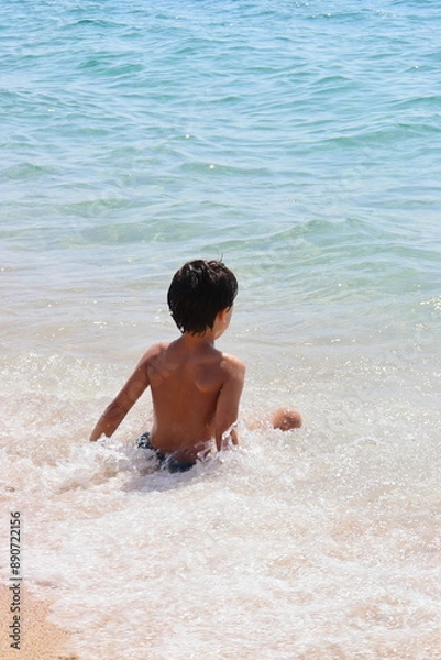 Fototapeta A boy is sitting in the ocean, looking out at the water in a beach.