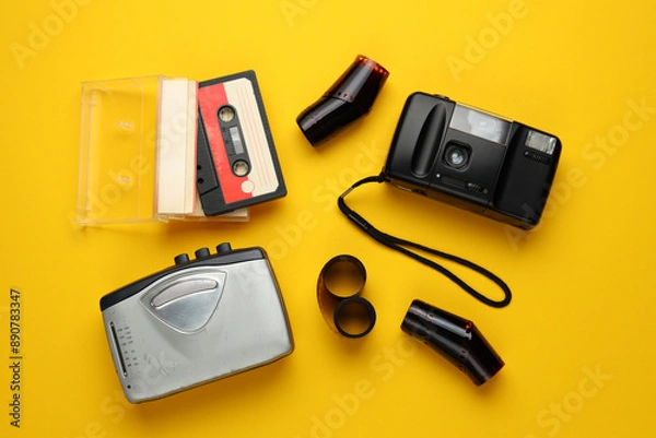 Fototapeta A collection of old-fashioned audio equipment and a camera. The camera is black and the equipment includes a cassette tape, a cassette tape player, and a camera