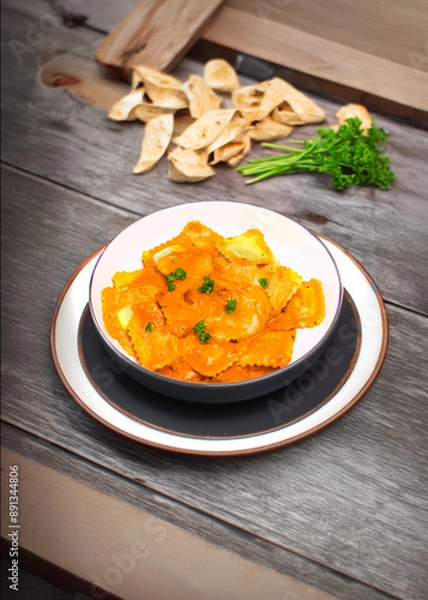 Obraz A delectable bowl of butternut squash ravioli in creamy sauce, garnished with fresh parsley. The rustic background includes bread crisps and a sprig of fresh parsley, enhancing the culinary appeal.
