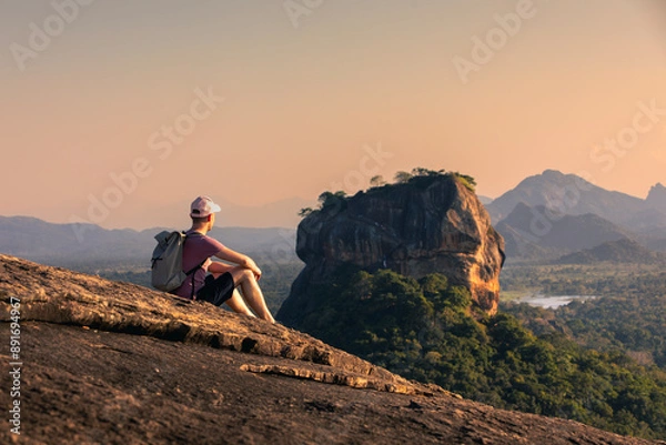 Fototapeta Man with backpack sitting on rock and looking at landscape at sunset. Beautiful scenery with Sigiriya rock. Solo traveler in Sri Lanka. .