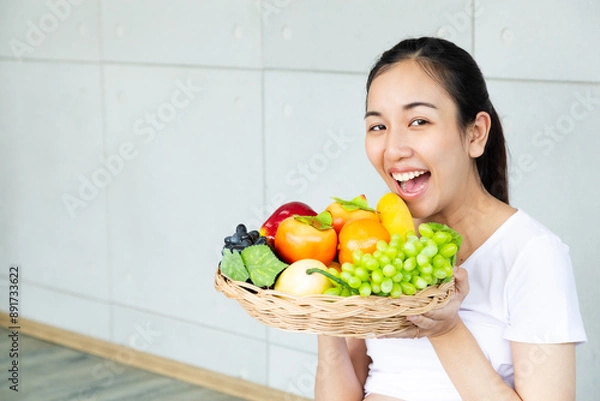 Fototapeta Young smiling woman posing with basket of fresh vegetables. Time of healthy food, bananas, apple, tomato, green grapes. Vegetables and Fruits.