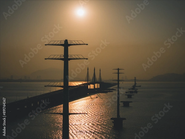 Fototapeta A stunning view of Penang Bridge at sunrise, framed by two TNB monopole electric transmission towers. The golden morning light casts a warm glow over the scene, highlighting the bridge's structure.