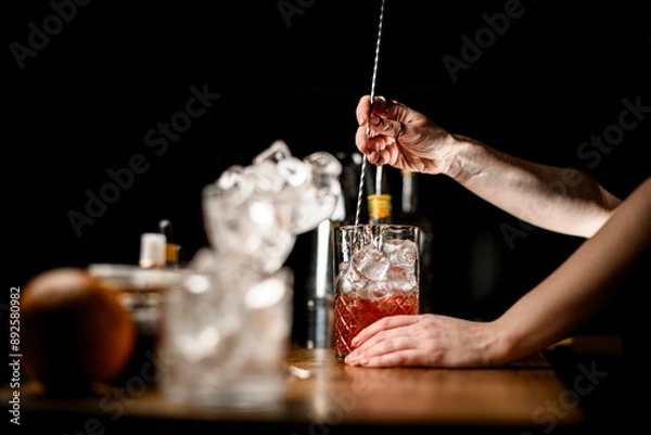 Fototapeta Focus on the background where female hands of a bartender stir a brown cocktail with ice