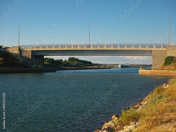 Fototapeta Bridge on the flowing river under the cloudy blue sky in Frontignan