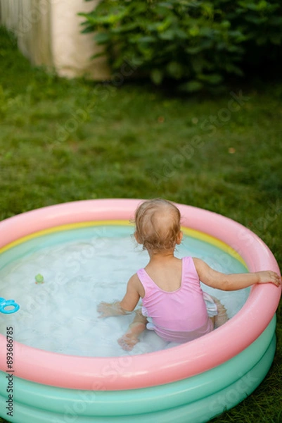 Fototapeta Baby in a pink swimsuit is swimming in inflatable pool in the backyard. No face. Back view. 
