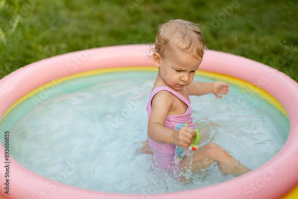 Fototapeta Baby in pink swimsuit is climbing out of an inflatable pool on the grass in the backyard. 