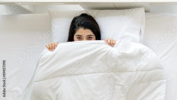 Fototapeta A woman is lying in bed, partly hidden under a white blanket, with a playful expression in her eye.