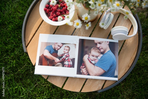 Fototapeta summer still life. Outdoor family photo album, headphones, daisies in a vase and raspberry berry on a plate lie on a table in the garden. family memories. happy summer holidays