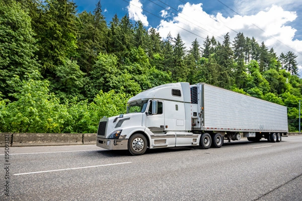 Obraz Popular comfortable big rig semi truck tractor transporting cargo in refrigerated semi trailer driving on the wide highway road with green forest on the side