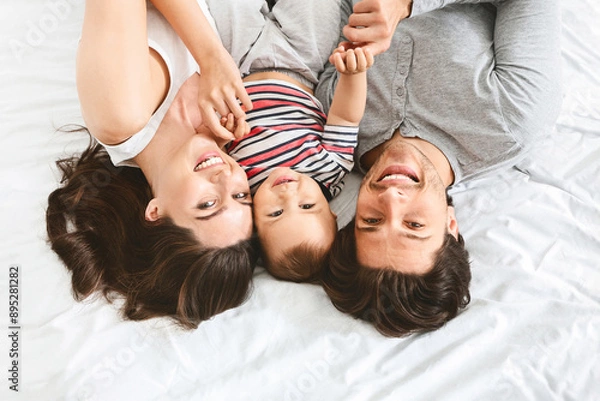 Fototapeta A happy family of three is lying on a bed together, holding hands. The parents are looking down at their child, who is smiling up at them. They are all wearing casual clothing, top view
