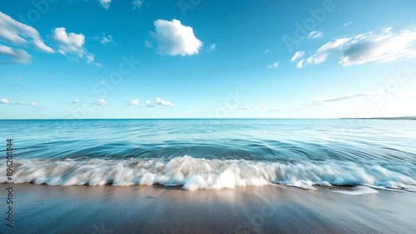 Fototapeta The calm ocean waves gently lap against the sandy shore under a clear blue sky.