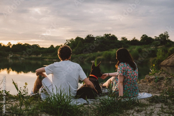 Fototapeta young happy couple having picnic and playing with their doberman dog outdoors near the lake during beautiful sunset