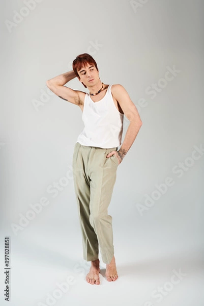 Fototapeta A stylish young man striking a pose in a white tank top and khaki pants against a grey studio background.