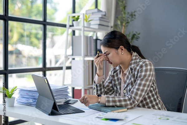 Fototapeta A woman is sitting at a desk with a laptop and a stack of papers