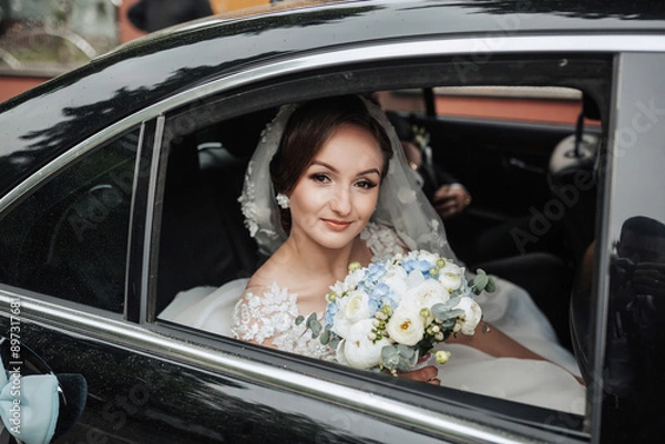 Fototapeta A bride is sitting in a car with a bouquet of flowers. She is smiling and looking out the window