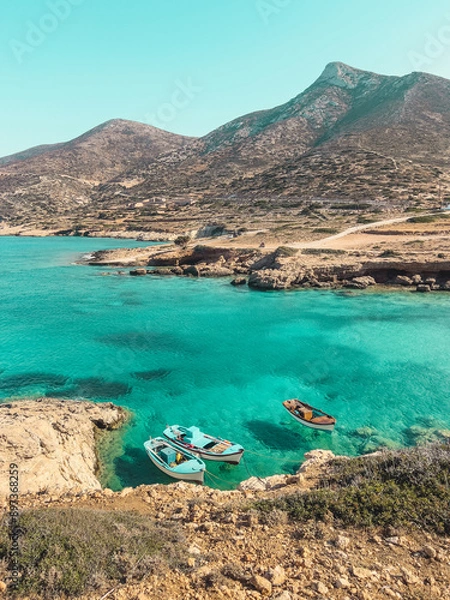 Obraz Three small boats gently float in the turquoise waters of a secluded cove in Donousa, Greece. Lush greenery contrasts with the brown cliffs in the distance