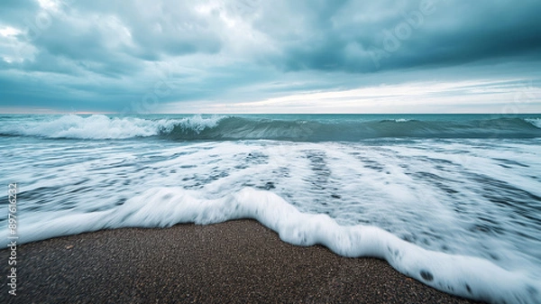 Fototapeta A serene view of waves crashing onto a sandy beach under a cloudy sky.