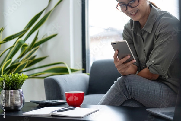 Fototapeta Businesswoman sitting at desk on couch in workplace or at home working on laptop and analyzing data on charts and graphs and writing on papers to make business plan and strategies for company, 