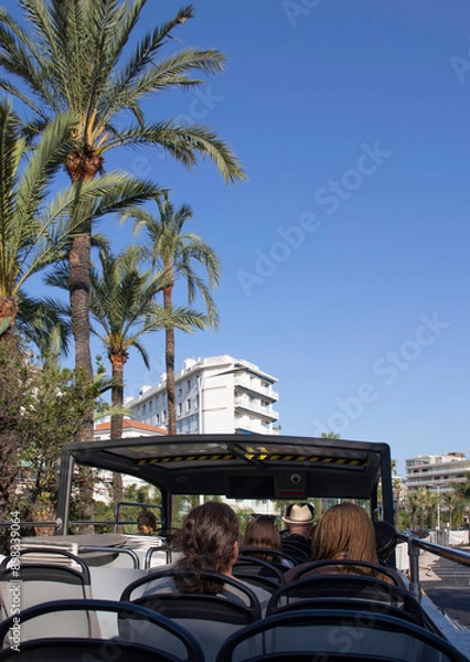 Fototapeta An open top tourist bus in Cannes in the south of France with a white building, blue sky and palm trees, from a tourist's point of view