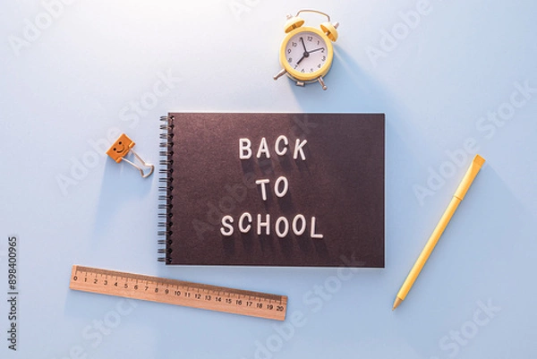 Fototapeta Black Notebook With The Words Back To School Written In White Letters On Blue Surface With Yellow Pencil, Ruler And Alarm Clock.