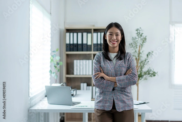 Fototapeta A woman is standing in front of a desk with a laptop and a stack of papers