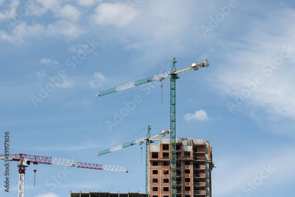 Fototapeta several construction cranes near an unfinished building against blue sky with light clouds