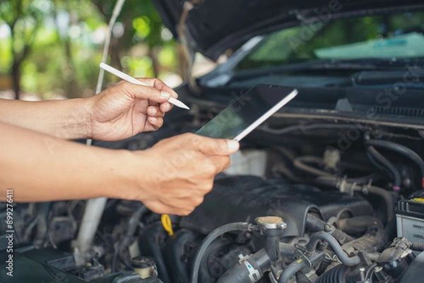 Fototapeta Close-up of Car mechanic noting repair parts during open car hood engine repair unrecognisable man wearing gray glove inspecting car engine and interior of hood of car.