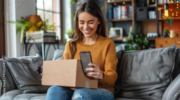 Fototapeta A smiling young woman is sitting on the sofa at home, holding a mobile phone and an open cardboard box in her hand, she is happy about the received package and the purchase.
