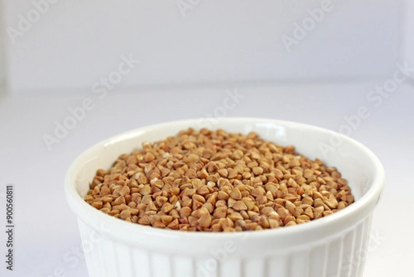 Fototapeta White bowl is filled with raw buckwheat groats, ready for cooking and serving