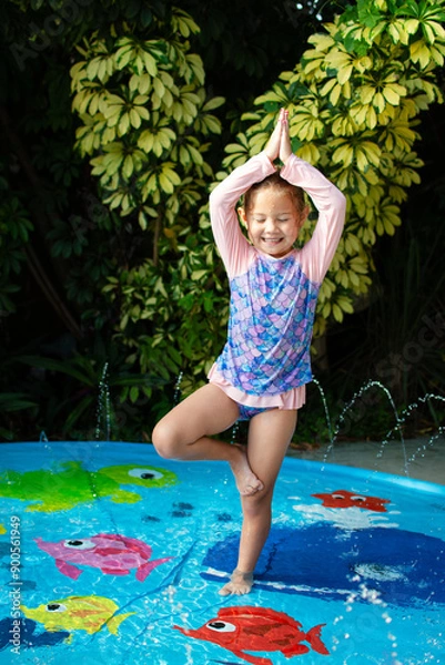 Fototapeta Cute Caucasian little girl doing yoga at the splash pad. Tree pose child. Summer fun kids concept