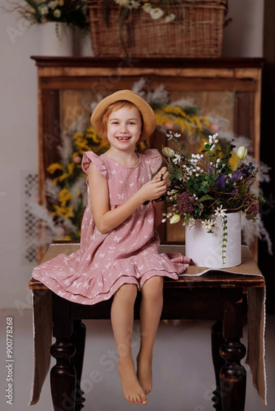 Fototapeta A young girl is sitting on a wooden table with a vase of flowers in front of her. She is wearing a pink dress and a straw hat. The scene is bright and cheerful