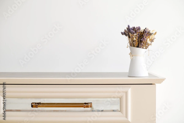 Obraz A bouquet of autumn asters in a white vase on a white chest of drawers.