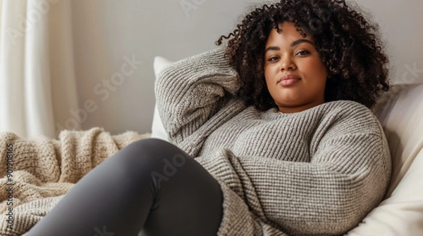 Fototapeta Chubby black woman wearing leggings and a sweater relaxing on the sofa, with her head resting in one hand. She has curly hair.  light gray background, relaxation concept 