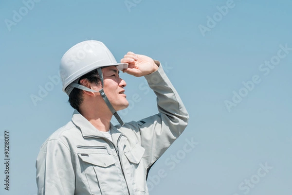Fototapeta 青空の下でヘルメットを被って空を見上げる作業着姿のアジア人男性作業員
