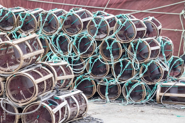 Fototapeta A large stack of cylindrical fishing traps is neatly arranged beside a waterfront dock, ready for use in capturing marine life, with boats and a town in the background.