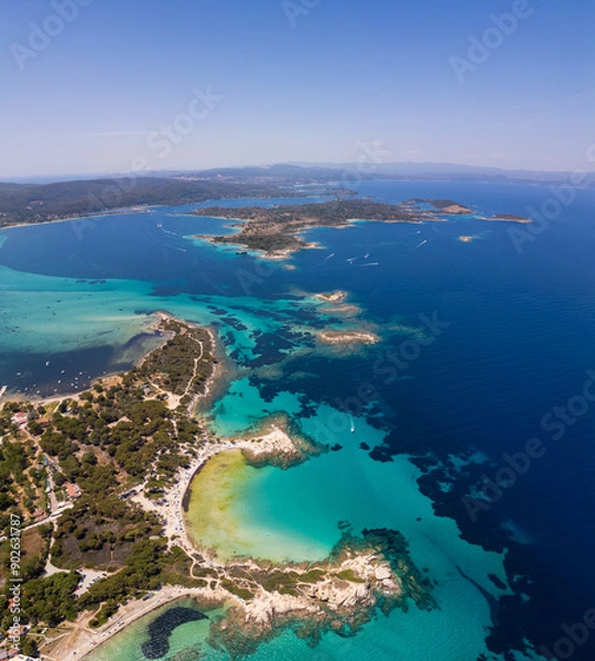 Fototapeta Aerial view of an island on Sithonia Peninsula, Greece, with beaches and rocks, surrounded by crystal-clear waters. Yachts and boats anchor in turquoise waters. Nearby islets and reefs visible