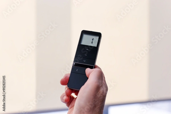 Obraz Remote control for window blinds. Man holds in his hand black remote control panel with display and operate motorized roller shades. Radio control electric blinds close-up.