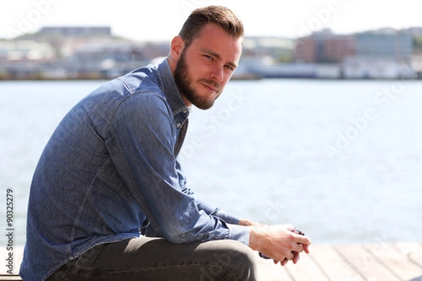 Fototapeta A handsome man sitting down outside wearing a jeans shirt with the river behind him on a sunny summer day.