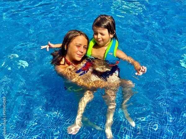 Obraz Two children happily swimming in a bright blue pool. They are enjoying the water, with one wearing a life jacket and both smiling.
