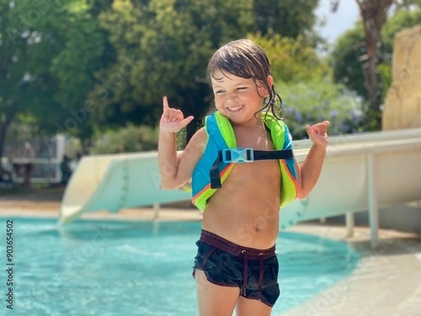Obraz A laughing 3-year-old Caucasian boy wearing a life vest, standing by the pool with horizontal copy space.
