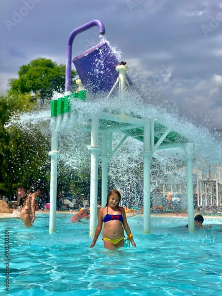 Obraz A 9-year-old Caucasian girl stands under a water bucket splash area, having fun at a pool on a sunny day.
