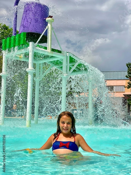 Obraz A close-up of a smiling 9-year-old Caucasian girl standing under a water bucket splash area, enjoying a sunny day at the pool.