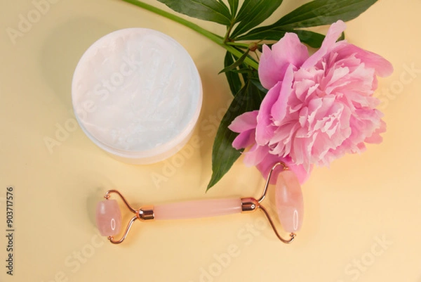 Fototapeta Massage with a quartz roller for the face with pink natural jade on a background of pink peonies. Guasha massage tool. Rejuvenation and lifting at home.