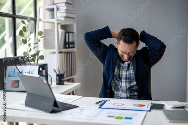 Fototapeta A man is sitting at a desk with a laptop and a stack of papers